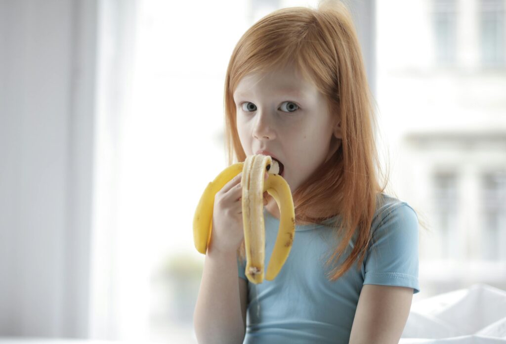 Flicka äter banan och ser frågande ut.